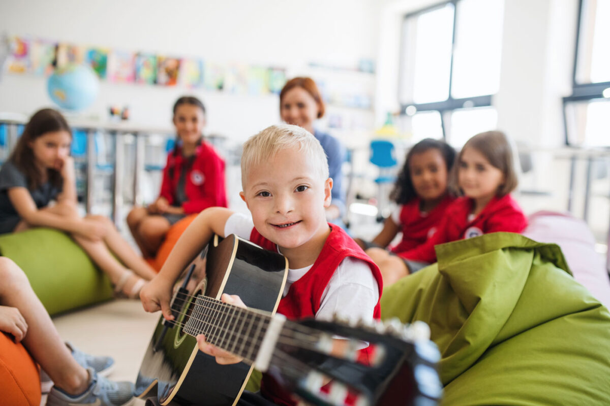 Un bambino che ride tenendo in mano una chitarra. Il bambino è seduto in un gruppo di molti bambini e un adulto.